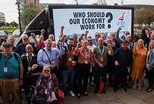 Workers in front of a billboard saying ' Who should our economy work for?'